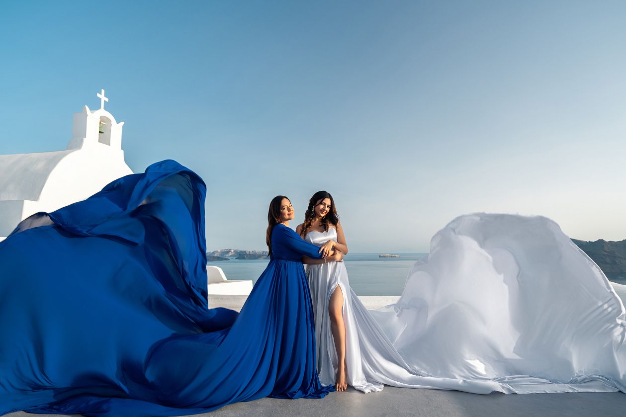 Mom and daughter share a tender embrace as their royal blue and white flying dresses billow dramatically around them against Santorini's whitewashed church and endless Aegean horizon.